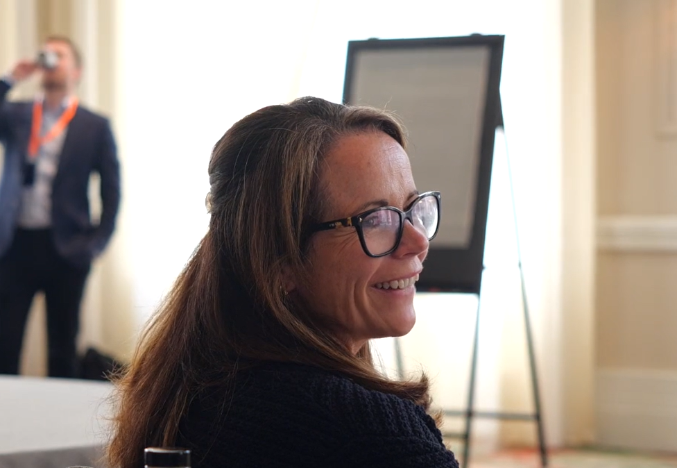A clinician smiling while seated at a round table during the Envisage Clinical Circle event, with two other attendees talking in the background.
